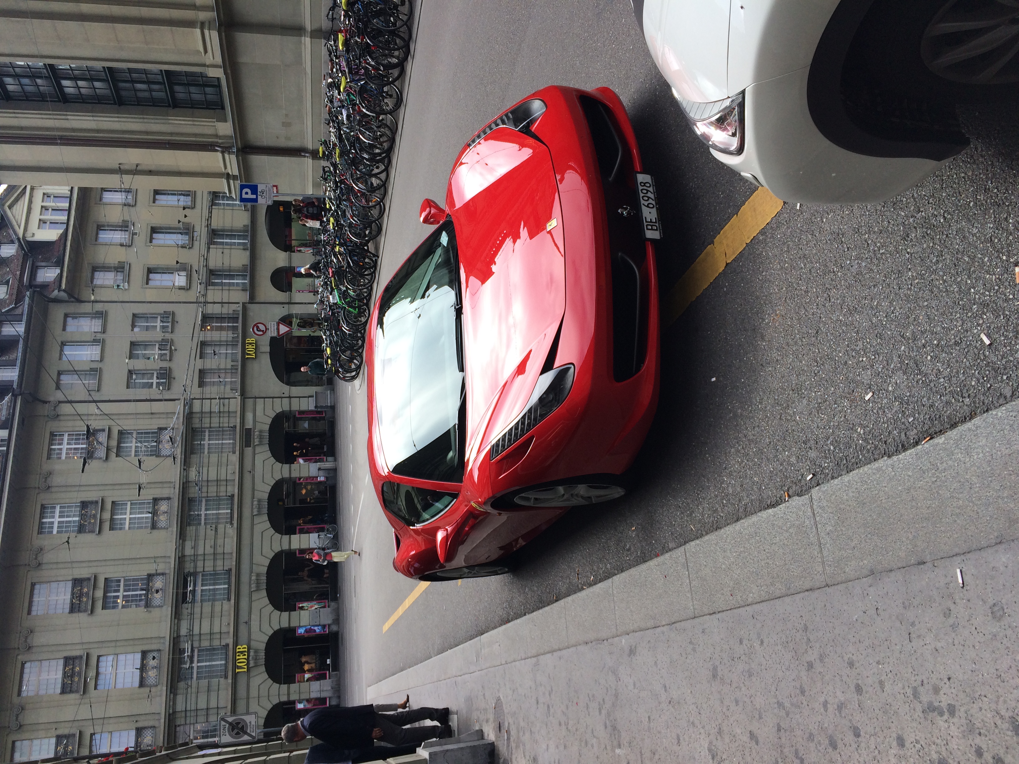 Czerwone Ferrari zaparkowane przy ulicy w centrum Berna, obok rzędu rowerów i arkad.