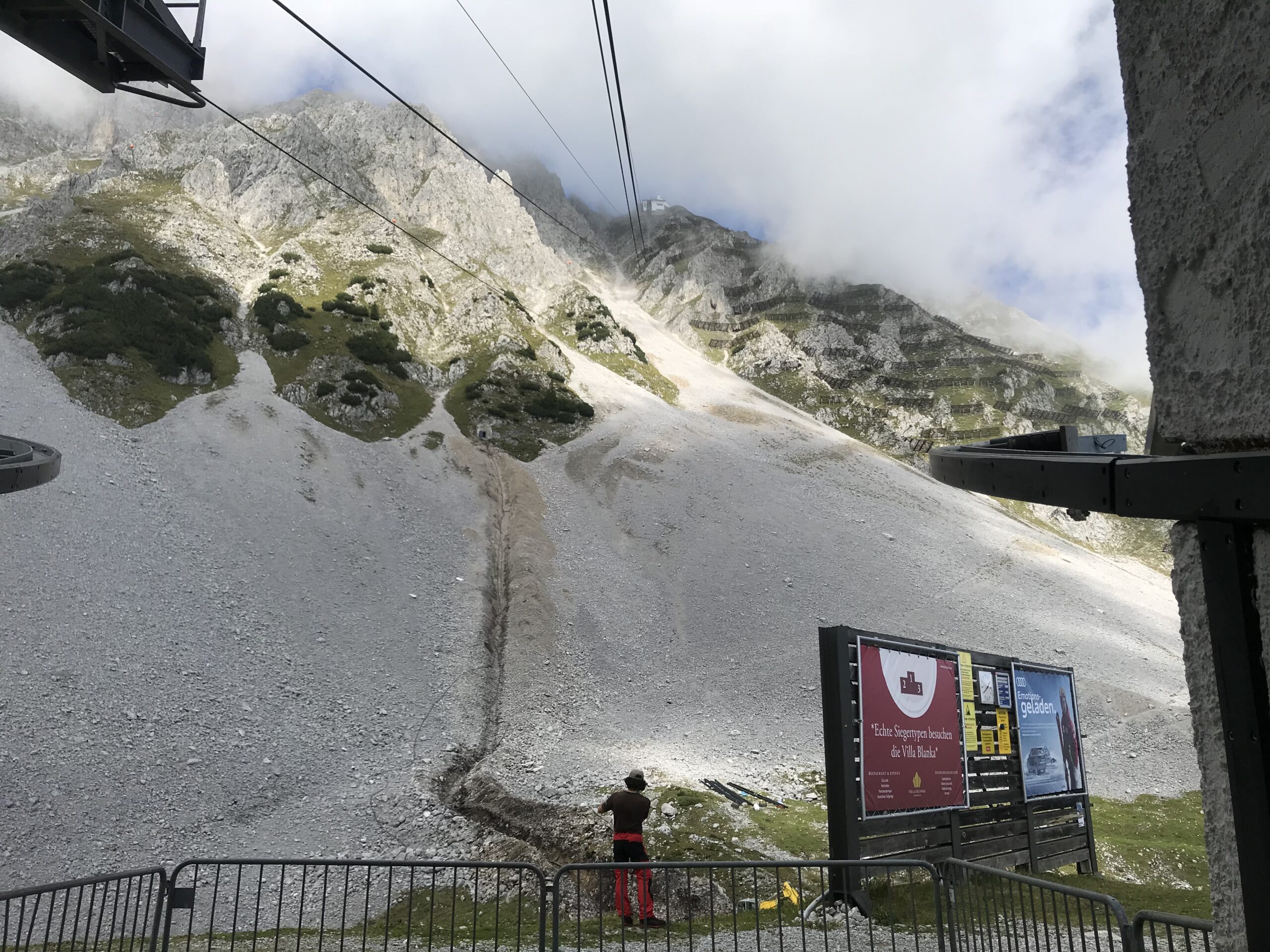 Kolejka Nordkette nad Innsbruckiem Trasa kolejki Nordkette nad skalistym zboczem w Alpach nad Innsbruckiem, z chmurami i tablicami informacyjnymi przy stacji