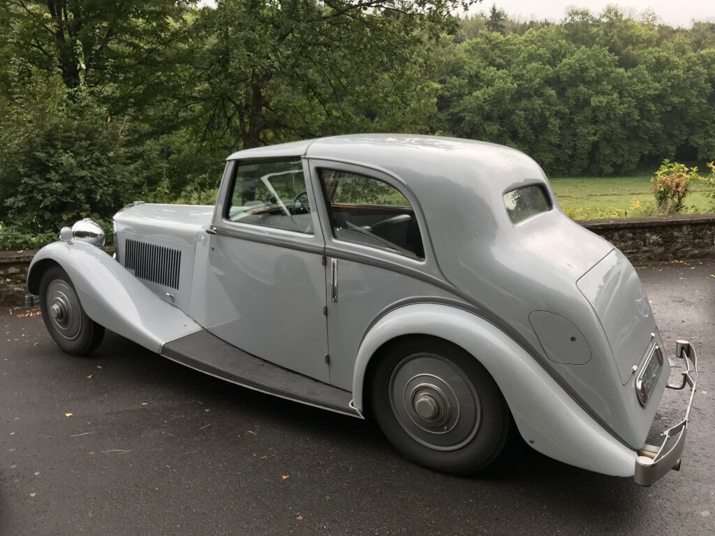 Bentley 4¼ Litre (Derby), Park Ward fixed-head coupé, ok. 1936–1939; srebrnoszary; widok z tyłu-lewego boku; osłony tylnych kół (spats), pojedyncze reflektory; zaparkowany na mokrym asfalcie, w tle zieleń i kamienny murek.
