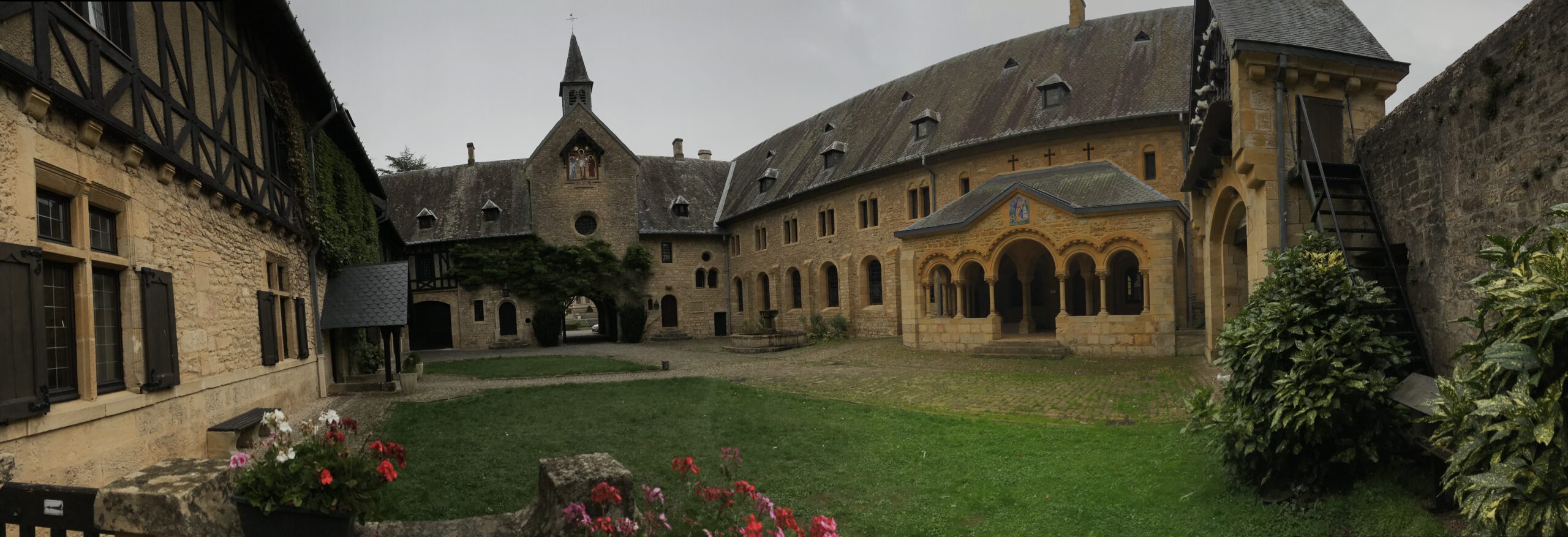Panorama dziedzińca opactwa Orval Panoramiczny widok dziedzińca opactwa Orval z kamiennymi budynkami, arkadowym krużgankiem, trawnikiem, kwiatami i pochmurnym niebem.