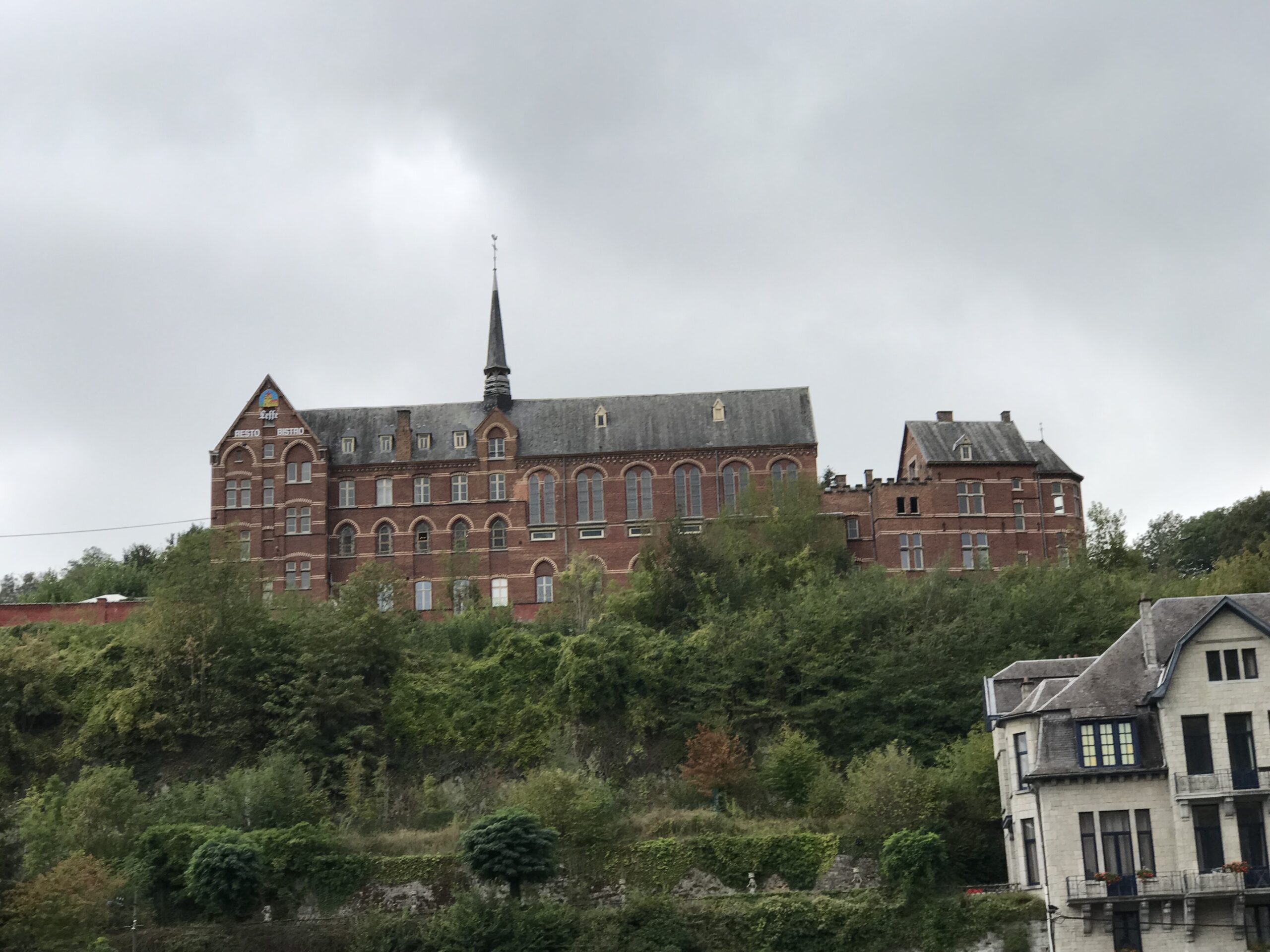 Zabudowania na wzgórzu nad Mozą w Dinant Ceglane zabudowania na wzgórzu nad Mozą w Dinant, widziane z przeciwnego brzegu rzeki pod pochmurnym niebem.