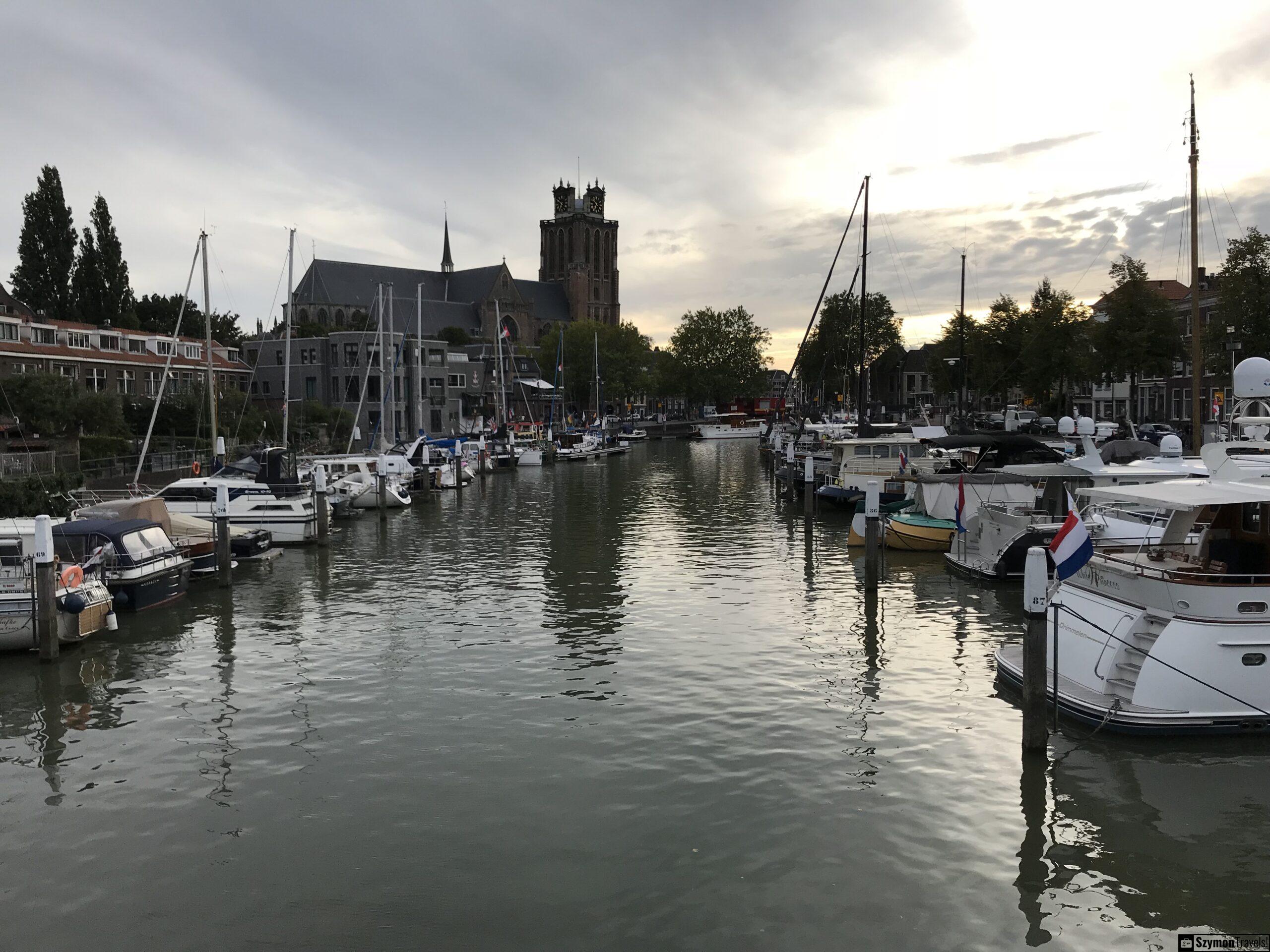 Stary port w Dordrecht z widokiem na Grote Kerk Stary port w Dordrecht z jachtami, łodziami mieszkalnymi, wodą oraz sylwetką Grote Kerk w tle pod pochmurnym niebem.