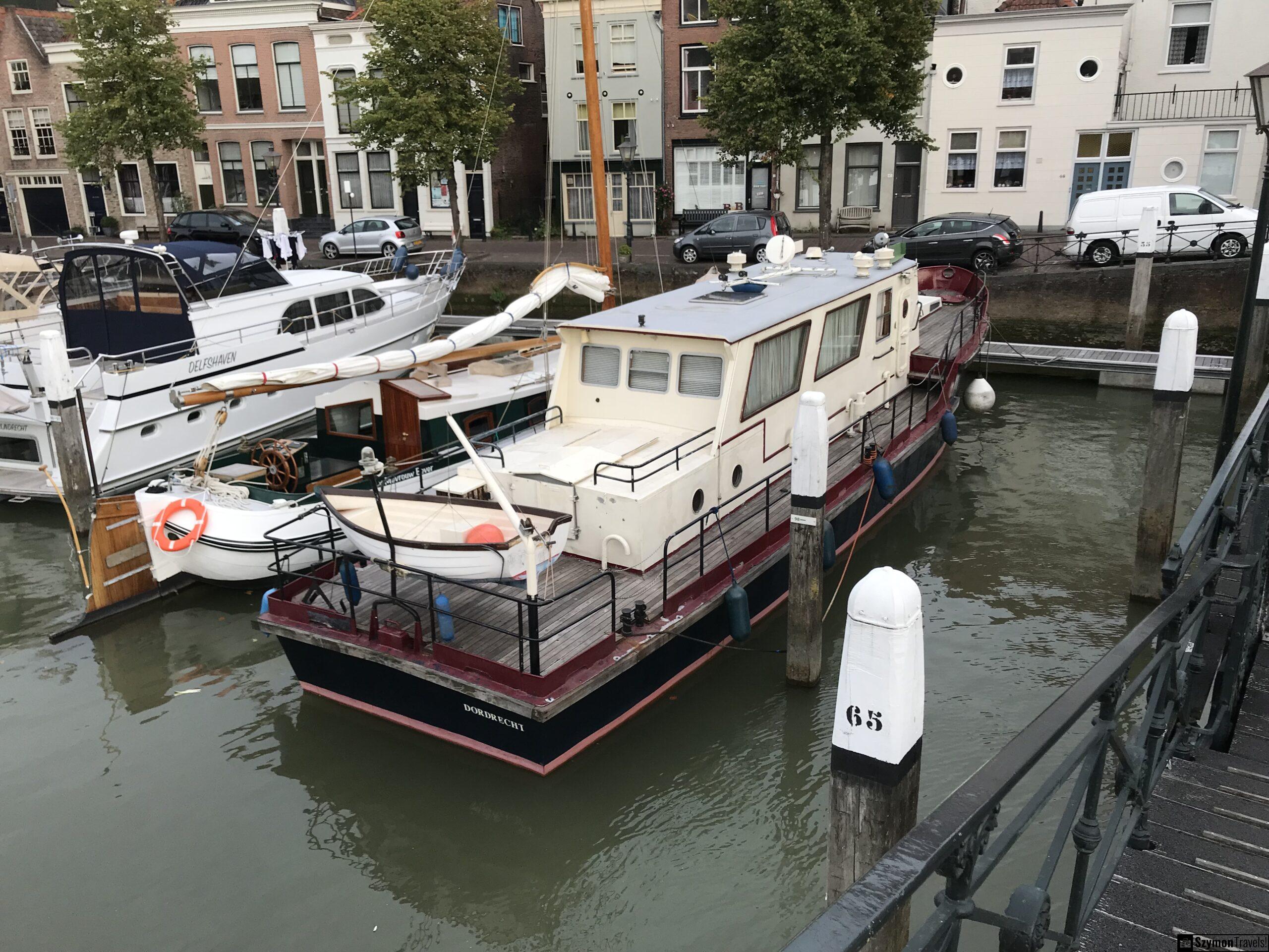 Łódź mieszkalna w starym porcie Dordrecht Duża łódź mieszkalna zacumowana w porcie Dordrecht, z białą boją, drewnianym pokładem, słupami cumowniczymi i kamienicami po drugiej stronie wody.