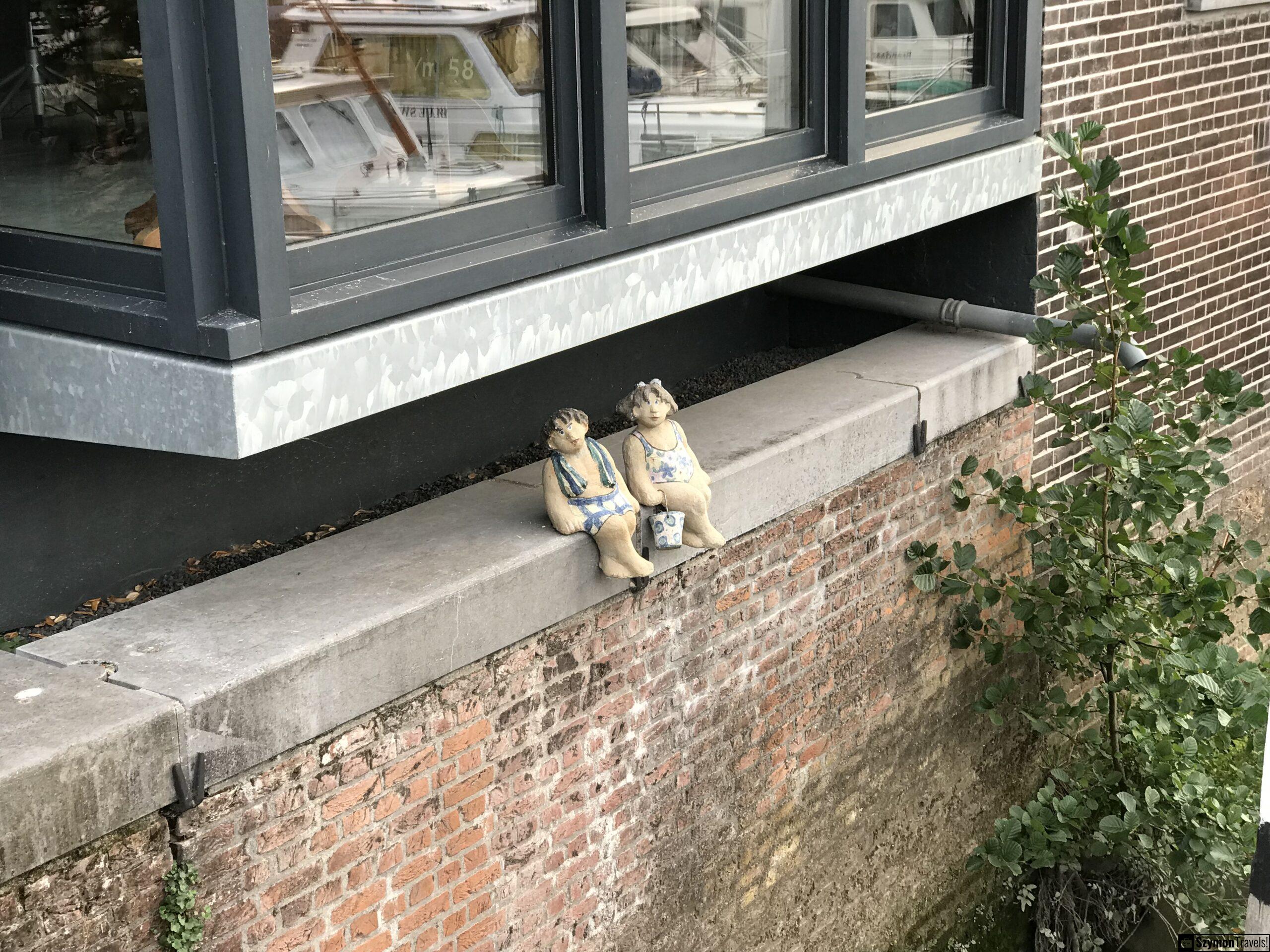 Ceramiczne figurki na murze w Dordrecht Dwie ceramiczne figurki siedzące na murze nad kanałem w Dordrecht, przy ceglanej ścianie i oknie odbijającym łodzie.