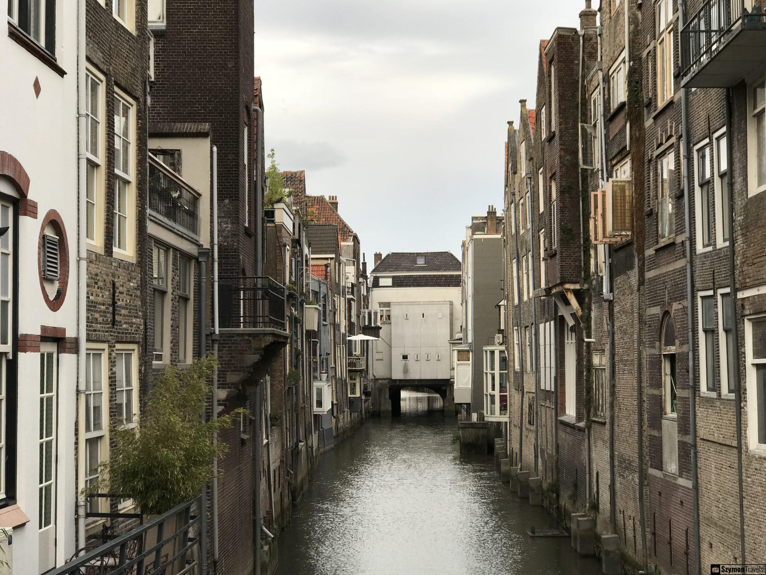 Kanał między kamienicami w Dordrecht Wąski kanał portowy w Dordrecht z wysokimi ceglanymi kamienicami po obu stronach i przejściem pod białym budynkiem na końcu.