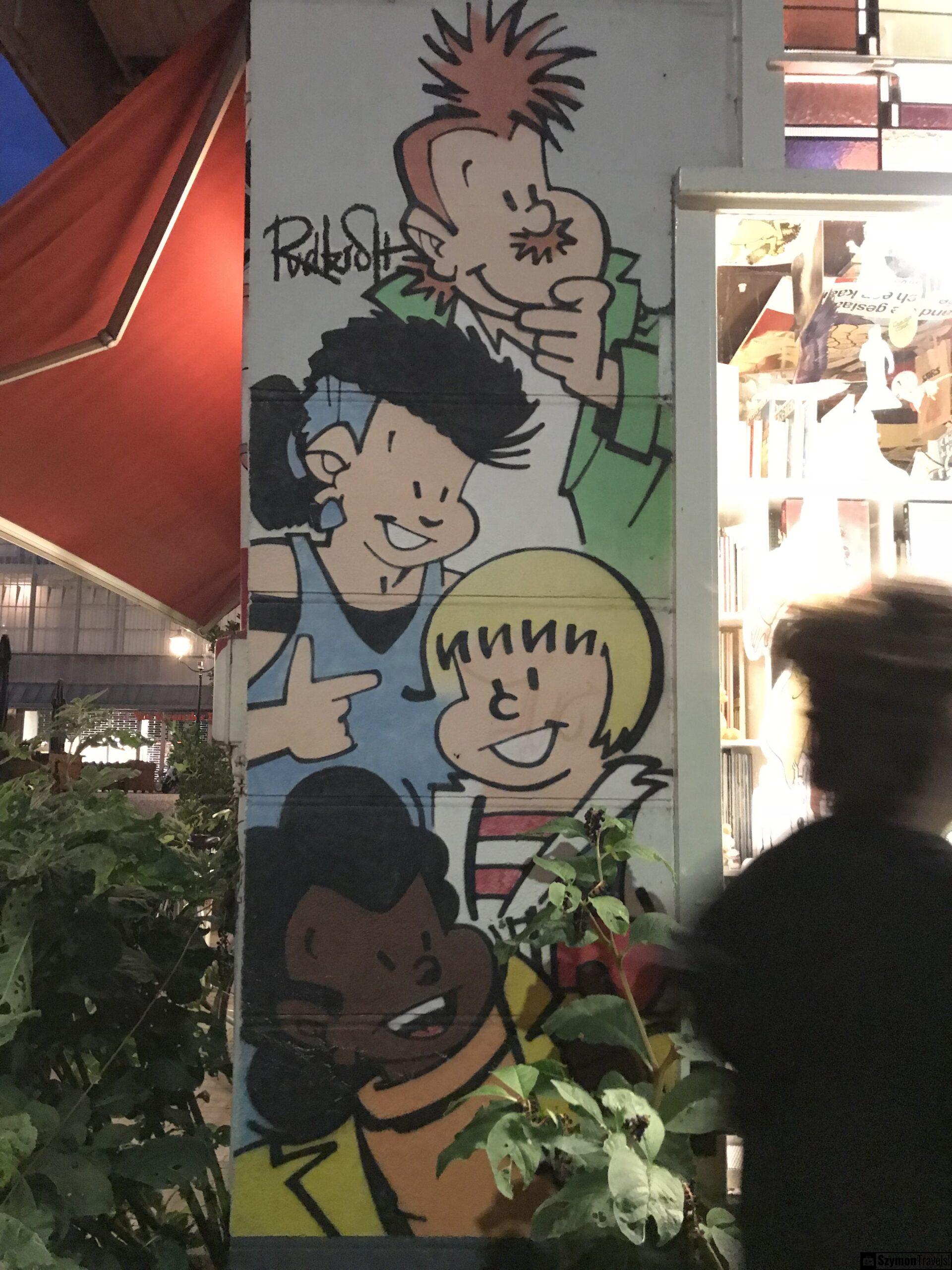 Kolorowy mural z postaciami w Dordrecht Kolorowy mural z rysunkowymi postaciami na ścianie w Dordrecht, sfotografowany wieczorem przy roślinach i przechodniach.