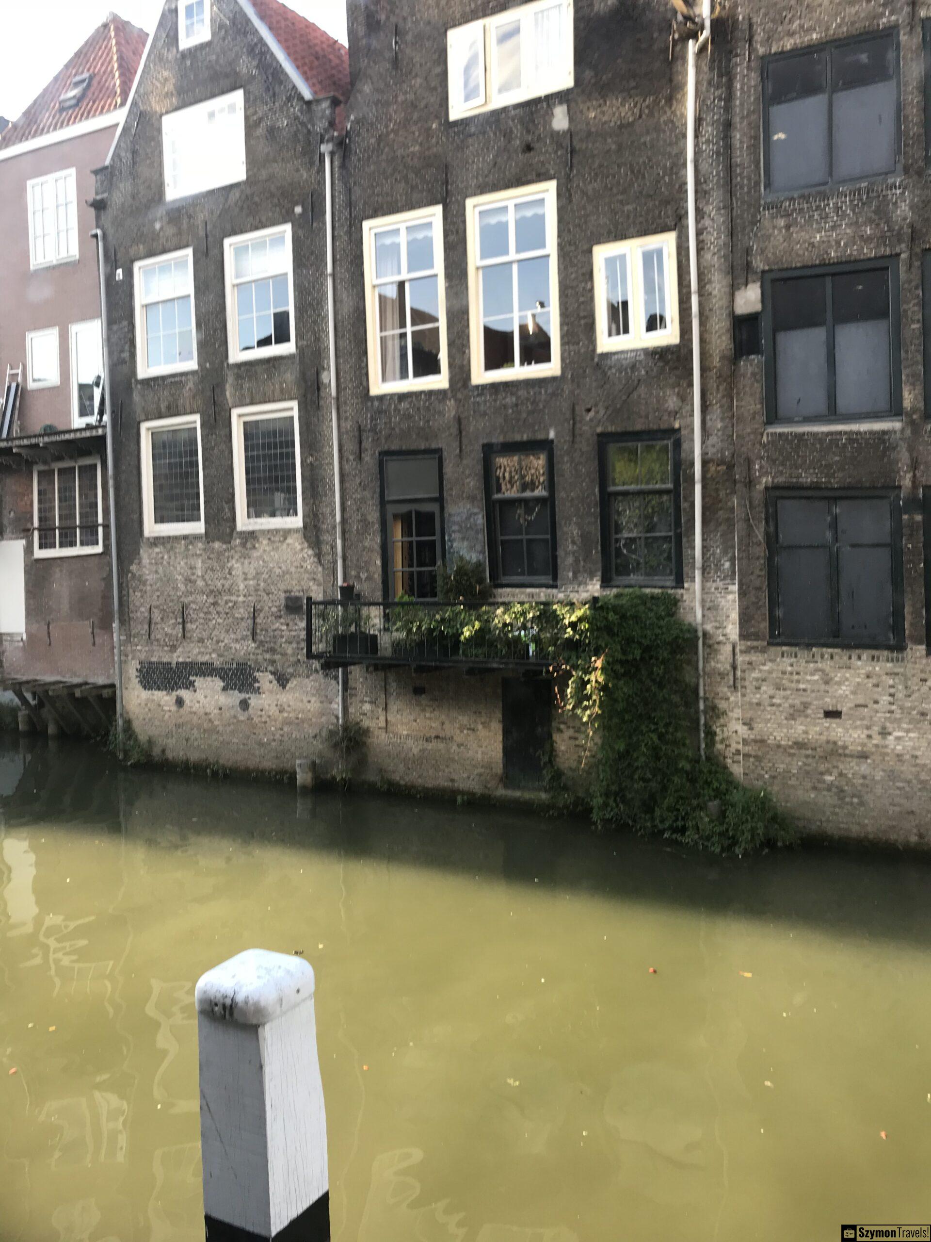 Ceglane domy nad kanałem w Dordrecht Stare ceglane domy stojące bezpośrednio nad kanałem w Dordrecht, z zielonkawą wodą i białym słupem cumowniczym na pierwszym planie.
