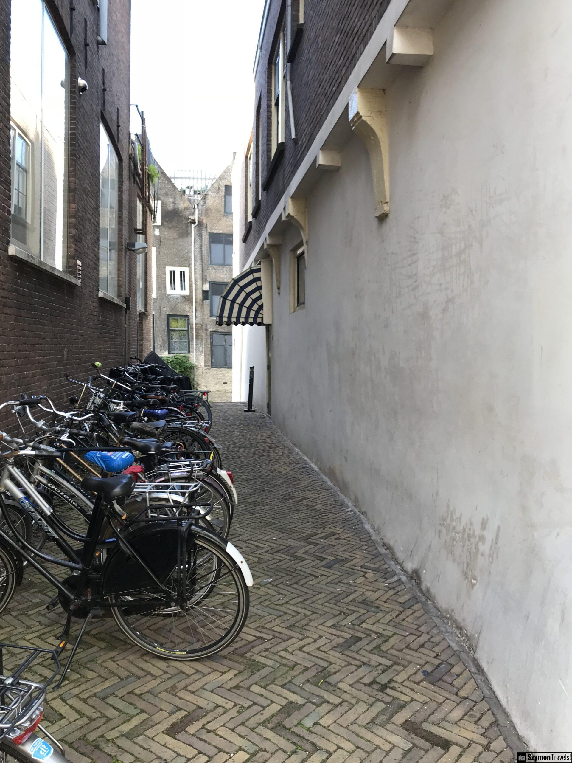 Wąska uliczka z rowerami w Dordrecht Wąskie przejście w Dordrecht z ceglaną nawierzchnią, zaparkowanymi rowerami po lewej stronie i pasiastym daszkiem nad wejściem.