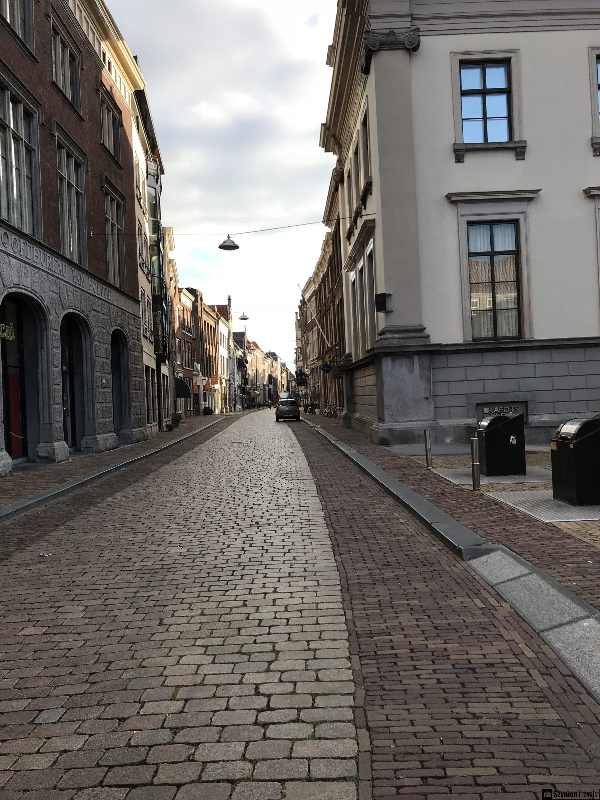 Brukowana ulica w centrum Dordrecht Brukowana ulica w centrum Dordrecht z historycznymi kamienicami po obu stronach, latarnią zawieszoną nad jezdnią i pojedynczym samochodem w oddali.