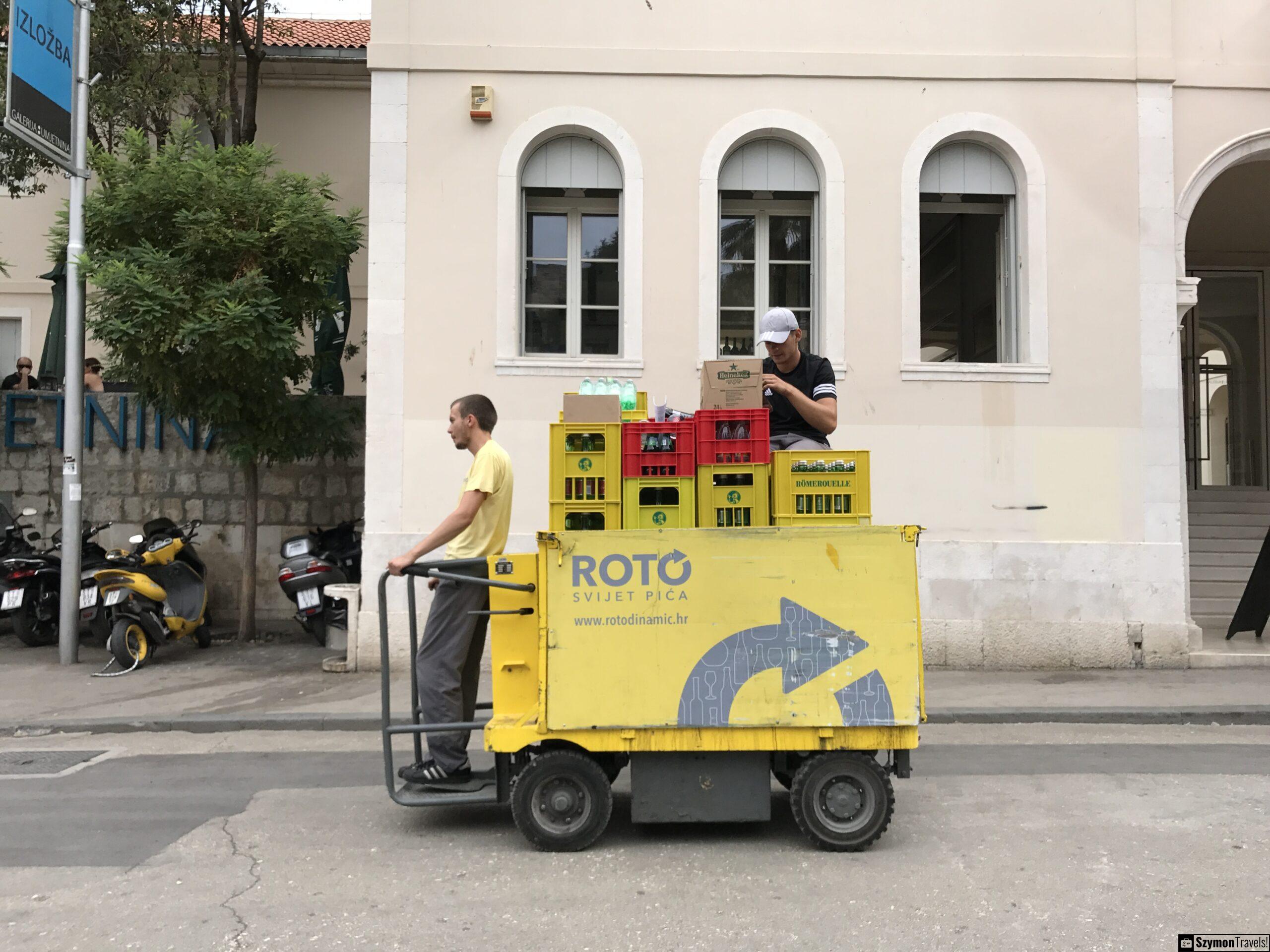 Dostawa napojów na ulicy w Splicie Żółty pojazd dostawczy z napojami na ulicy w Splicie, z pracownikami rozwożącymi skrzynki z wodą i napojami.