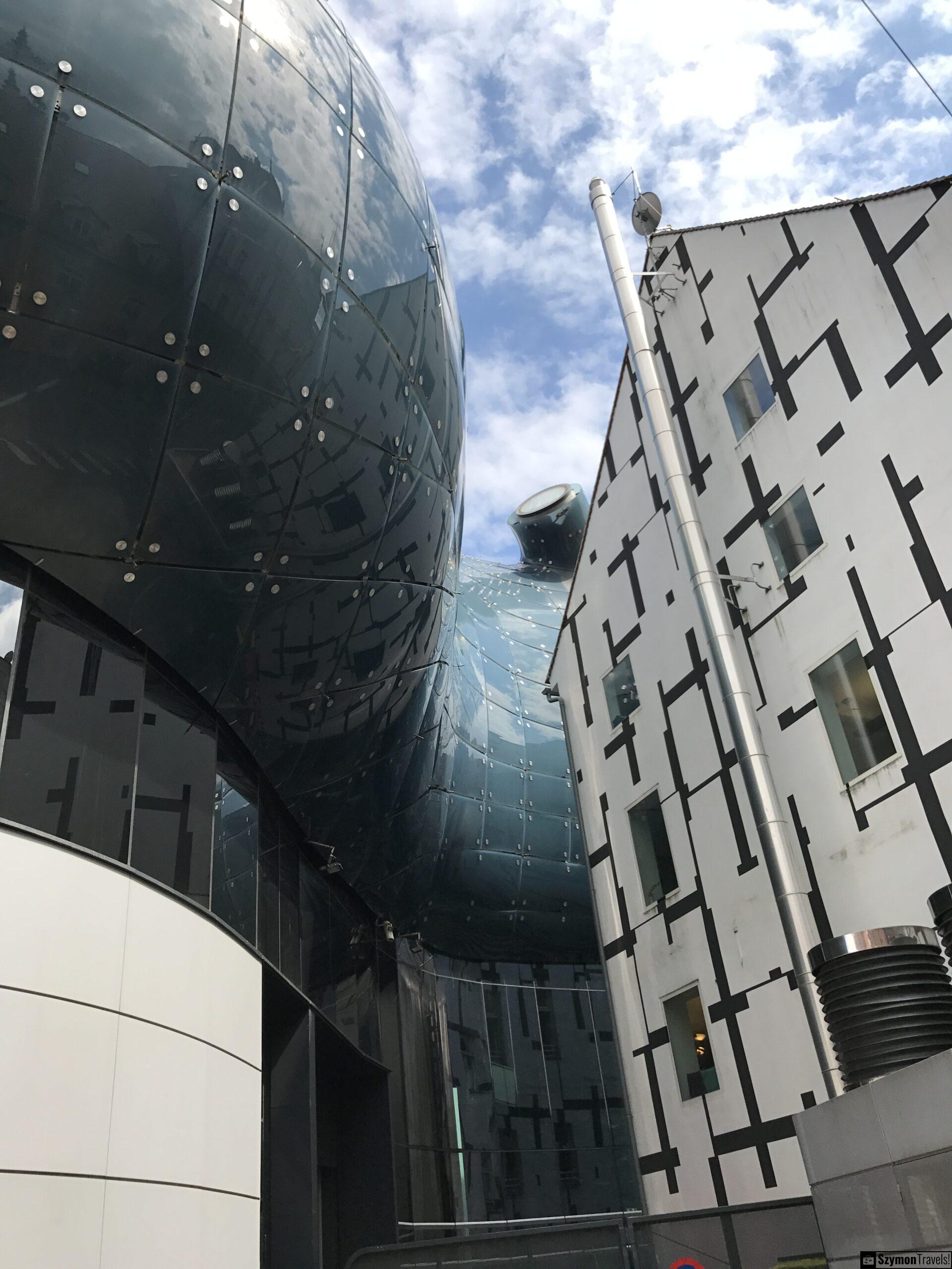 Kunsthaus Graz z bliska Zbliżenie na błyszczącą, biomorficzną fasadę Kunsthaus Graz i sąsiedni biały budynek z czarnym wzorem.