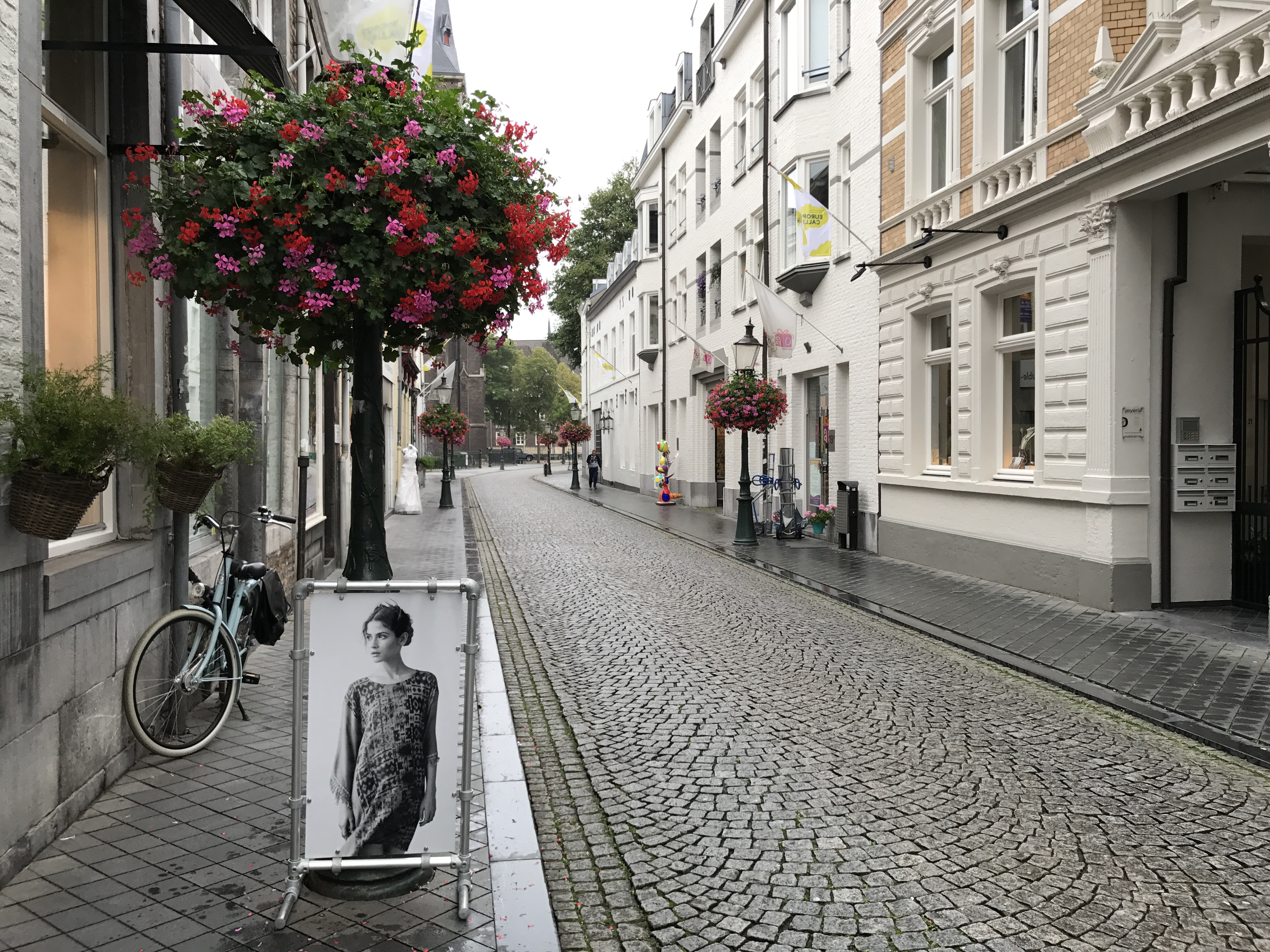 Spokojna brukowana ulica w Maastricht z białymi fasadami, wiszącymi kwiatami, rowerem i szyldem przed sklepem.