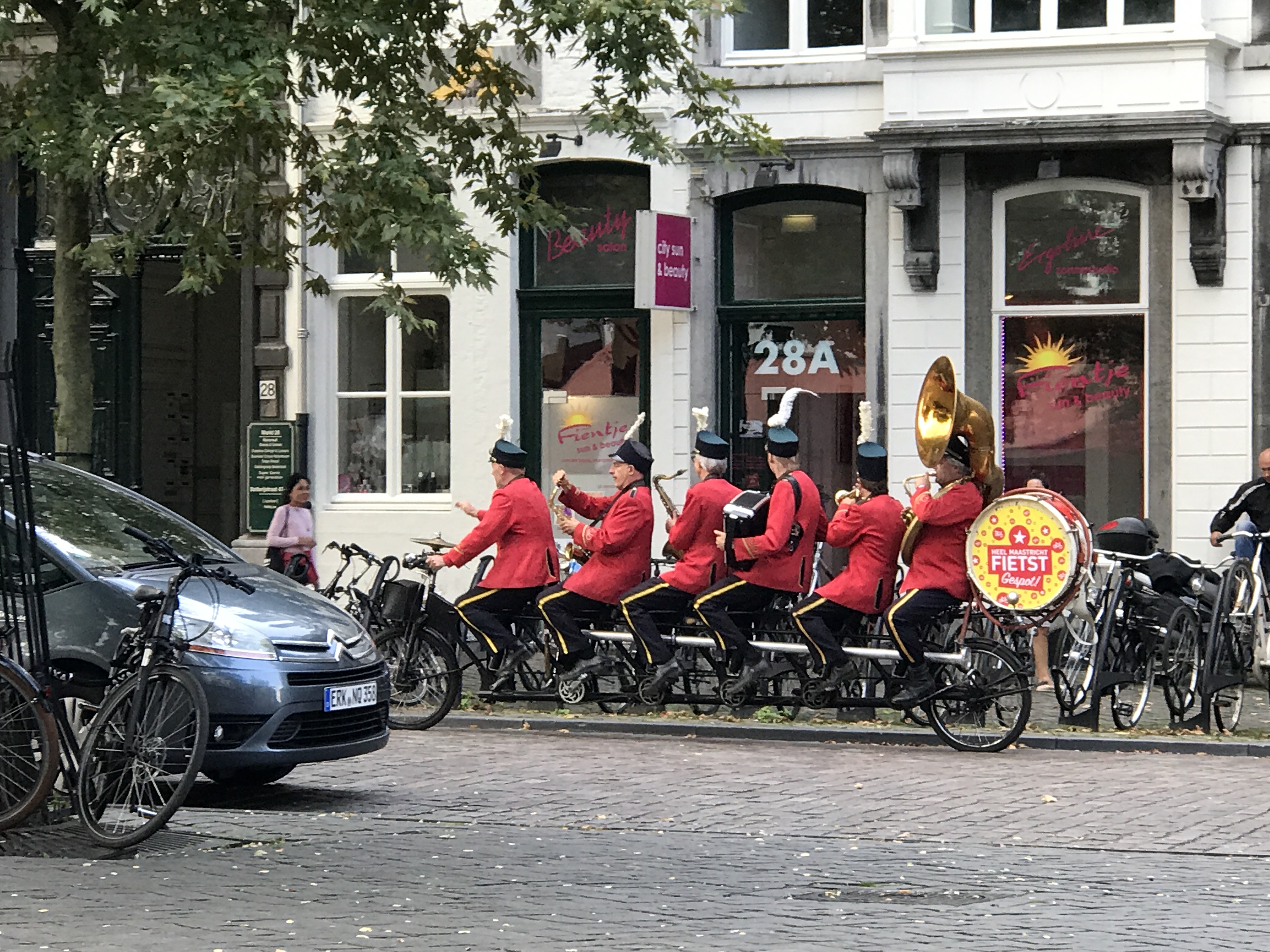 Rowerowa orkiestra w Maastricht, muzycy w czerwonych kurtkach jadący na wspólnym wieloosobowym rowerze i grający na instrumentach.