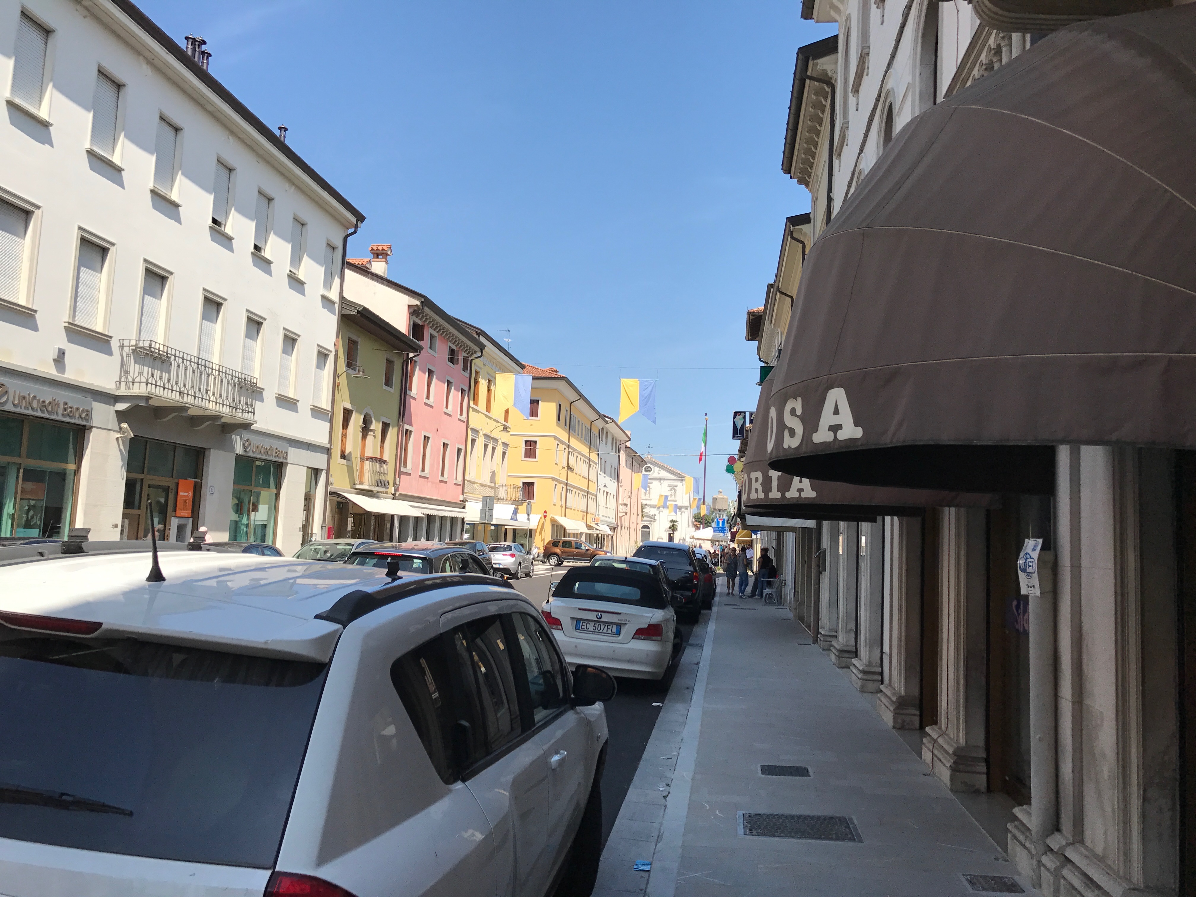 Ulica w Palmanovie z kolorowymi kamienicami, samochodami i chodnikiem pod arkadami