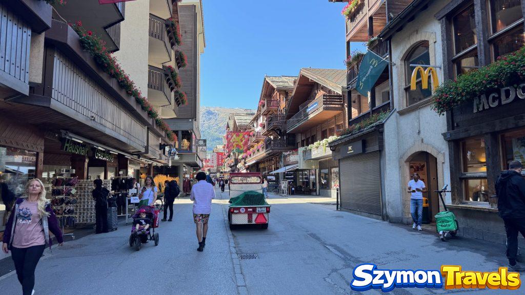 Ulica w Zermatt bez prywatnych samochodów Główna ulica w Zermatt z drewnianymi balkonami, sklepami, pieszymi, małym pojazdem elektrycznym i górami w tle.