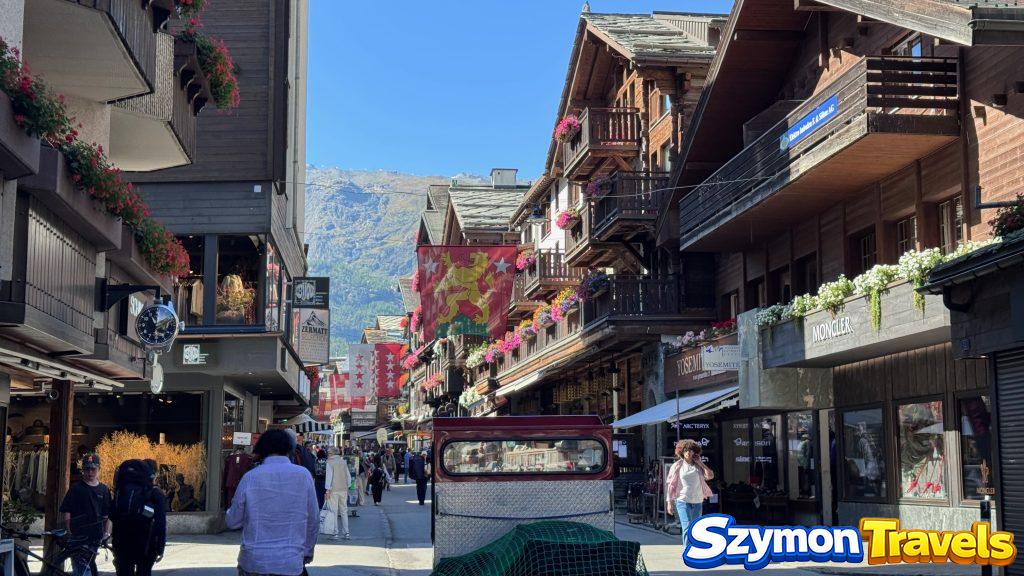 Ulica handlowa w Zermatt z drewnianymi balkonami, flagami szwajcarskimi, sklepami i górskim tłem.