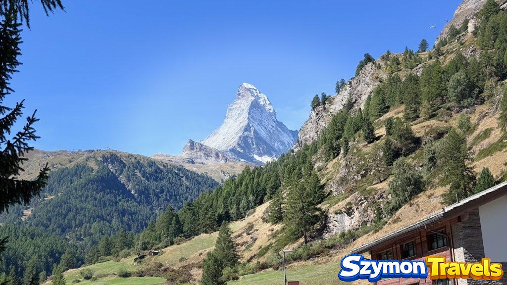 Matterhorn widoczny między zalesionymi zboczami i alpejską zabudową w okolicach Zermatt.