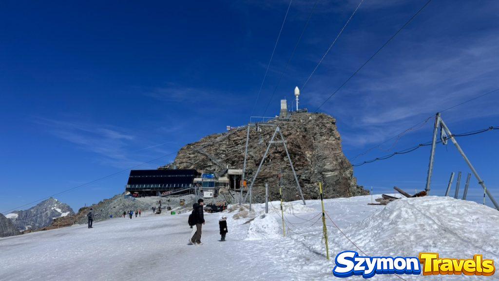Śnieżny teren przy Matterhorn Glacier Paradise, z budynkiem stacji, skałą, turystami i błękitnym niebem.