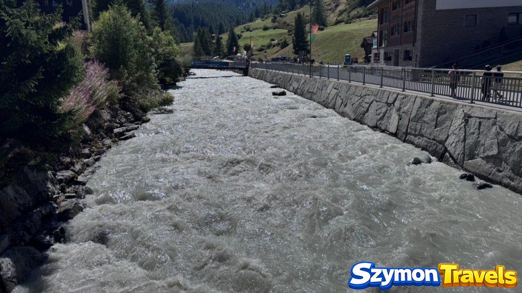Rwący górski potok w Zermatt, ujęty kamiennymi murami i chodnikiem wzdłuż brzegu.