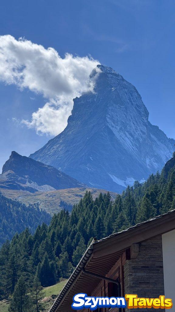 Matterhorn z chmurą nad Zermatt Matterhorn z chmurą opływającą szczyt, widziany ponad lasem i dachem alpejskiego budynku w Zermatt.