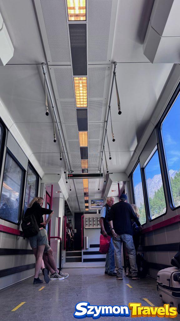 Wnętrze pociągu Zermatt Shuttle z Täsch do Zermatt, z pasażerami, miejscem na bagaże i dużymi oknami.