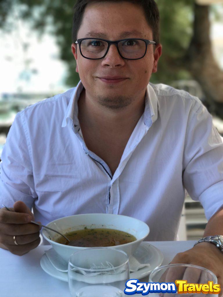 Autor siedzący przy stoliku w restauracji w Primoštenie, w białej koszuli, z głębokim talerzem zupy lub dania obiadowego przed sobą.