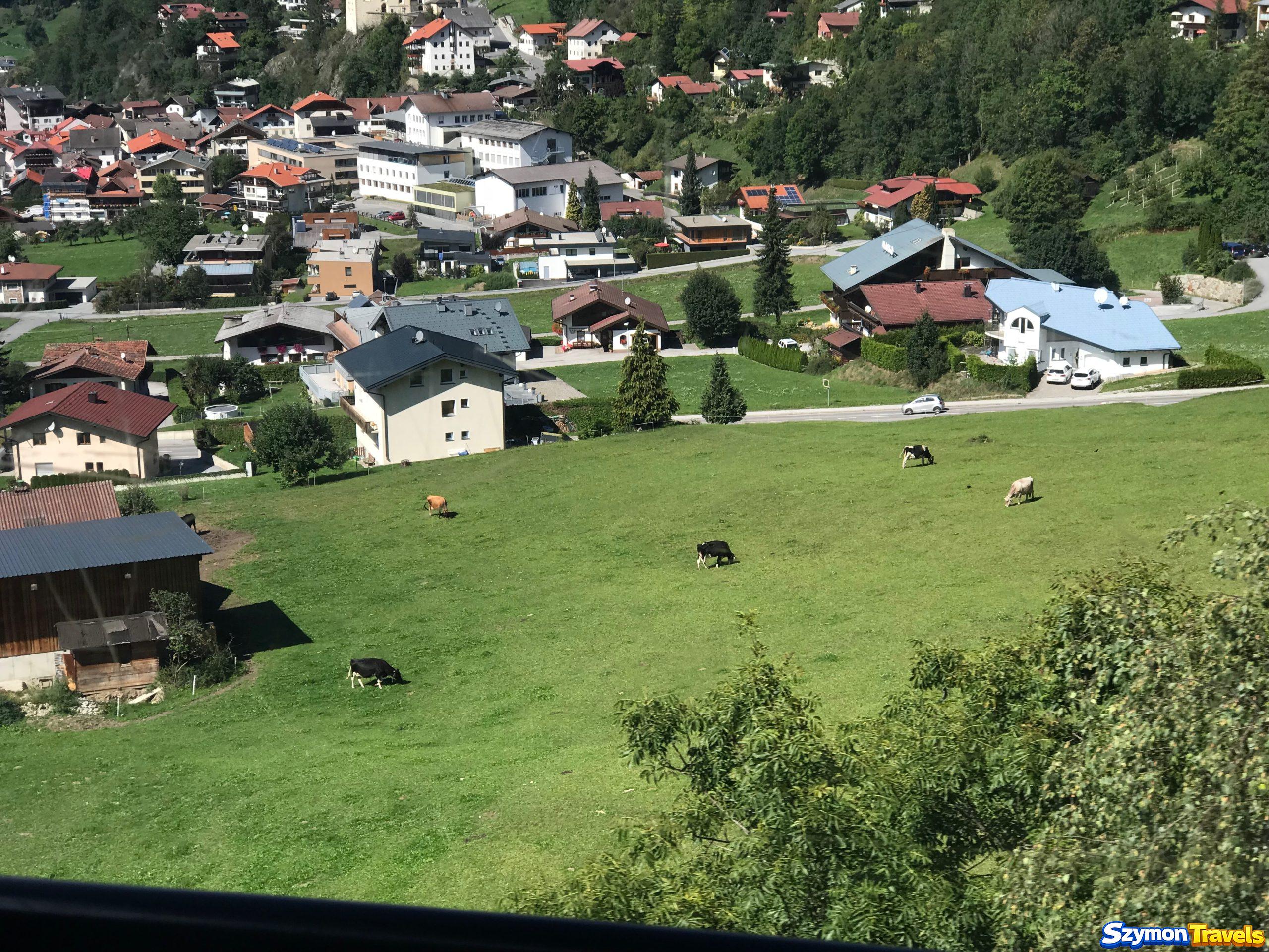 Pastwiska i Oetz widziane z gondoli Widok z gondoli nad Oetz na zielone pastwisko z krowami, domy miejscowości, drogi i lasy na zboczach.
