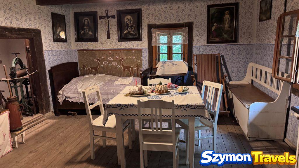 Izba z łóżkiem, stołem, świętymi obrazami i białymi krzesłami w Muzeum Wsi Opolskiej