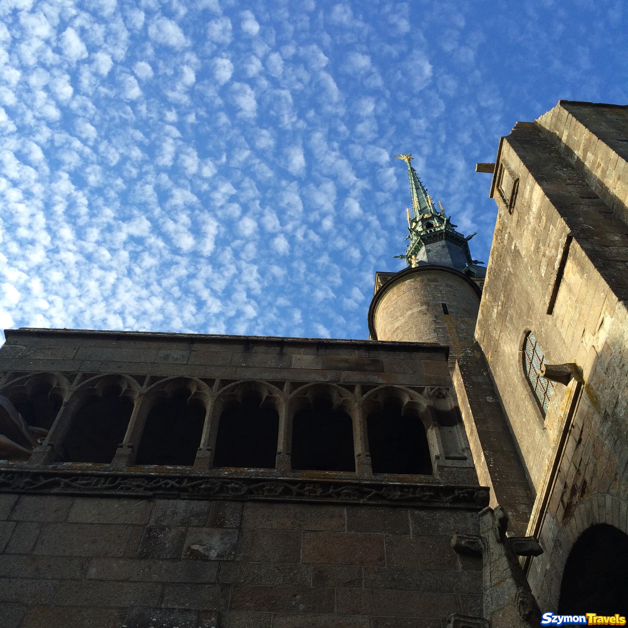 Wieża opactwa Mont-Saint-Michel i arkadowa galeria widziane od dołu na tle drobnych chmur