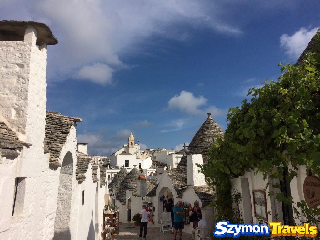 Uliczka Rione Monti w Alberobello z białymi trulli, sklepikami i widoczną w oddali wieżą kościelną