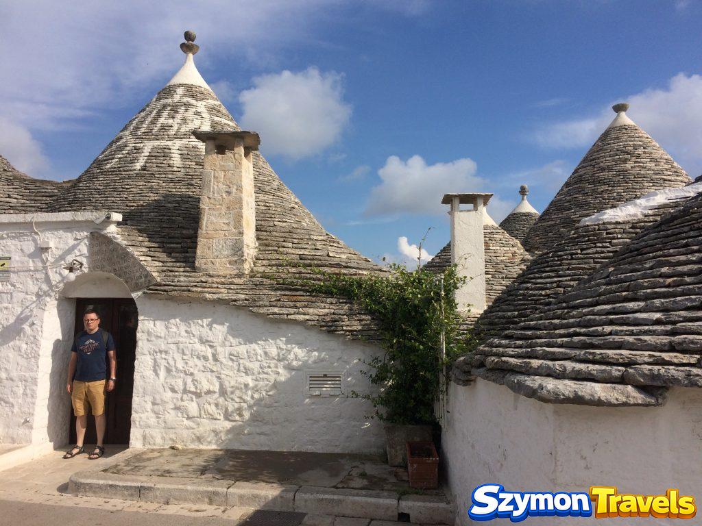 Szymon przed wejściem do zabytkowego trullo w Alberobello, otoczonego kilkoma stożkowymi dachami i białymi ścianami