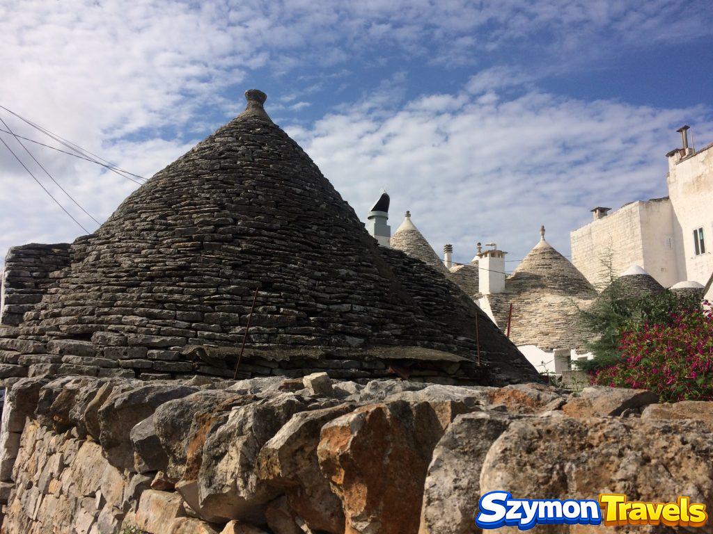 Kamienny dach trullo oglądany zza muru, z kolejnymi stożkami w tle w Alberobello
