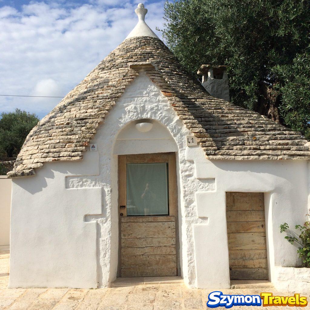 Bielony dom trullo z drewnianymi drzwiami, kamiennym stożkowym dachem i drzewem oliwnym w Alberobello